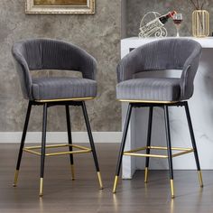 two grey velvet bar stools with gold trim and black metal legs in front of a marble wall