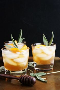 two glasses filled with ice and orange slices on top of a wooden table next to an olive branch