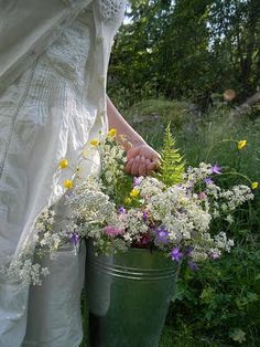 a woman in white dress holding a green bucket filled with wildflowers and daisies