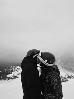 two people standing next to each other on top of a snow covered slope with mountains in the background