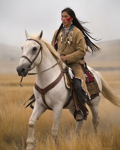 a native american woman riding on the back of a white horse in an open field