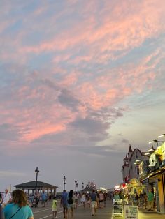 people walking on the boardwalk at dusk with pink clouds in the sky over buildings and shops