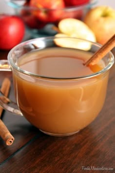 a glass cup filled with liquid and cinnamon sticks on top of a wooden table next to apples