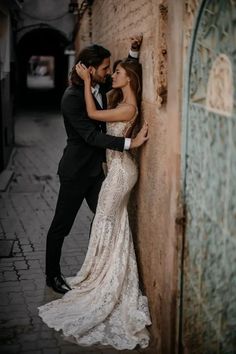 a bride and groom leaning against a wall in an alleyway, embracing each other