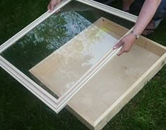 a person holding up a wooden frame with trees reflected in it on the grass,