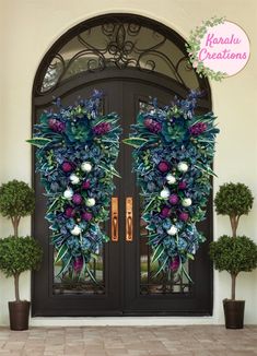 two large wreaths on the front door of a house with potted trees and bushes