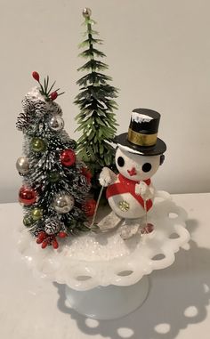 a small snowman next to a christmas tree on a white plate with silver ornaments