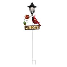 a red bird sitting on top of a welcome sign next to a lamp post with flowers