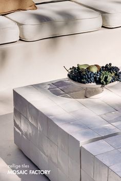 a bowl of fruit sitting on top of a white table next to a couch with pillows