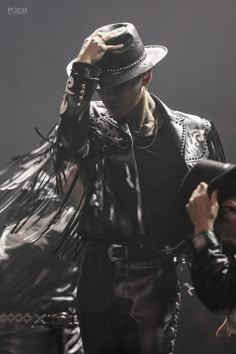 a man in a hat and leather outfit holding his hands up to his head while standing next to another man