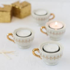 four tea cups with candles in them sitting on a white tablecloth next to a gold gift box