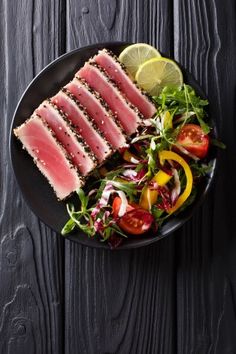 a black plate topped with meat, salad and lemon wedges on top of a wooden table