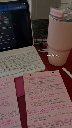 a laptop computer sitting on top of a desk next to a pink cup and pen