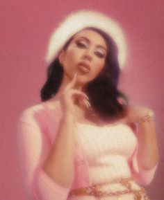 a woman wearing a white hat and holding her hand to her mouth while standing in front of a pink wall