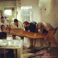 a group of people standing around a large fish in a kitchen next to a sink