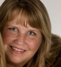 a woman with long blonde hair and blue eyes smiling at the camera while wearing a brown jacket