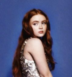 a woman with long red hair wearing a silver dress and posing in front of a blue background