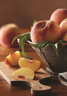 some peaches are sitting in a colander on a wooden table next to a cutting board