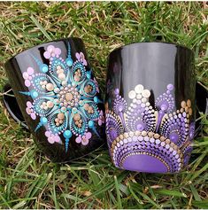 two coffee mugs sitting in the grass with designs painted on them, one is purple and the other is black