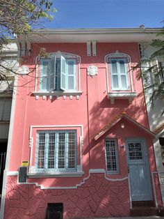 a pink house with white windows and shutters on the front, next to a tree