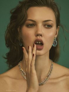 a woman with her hands to her mouth and wearing jewelry on her chest, standing in front of a green background