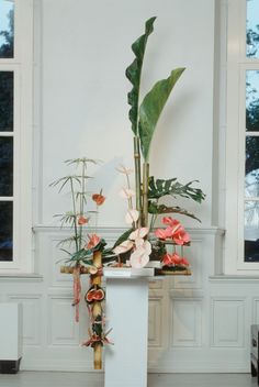 an arrangement of flowers and greenery on a pedestal in front of a wall with windows