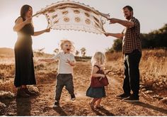 a family holding an umbrella in the middle of a dirt road with their toddler running towards them