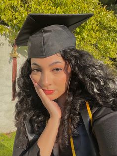 a woman wearing a graduation cap and gown with long curly hair sitting in front of a tree