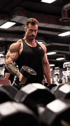 a man in black shirt holding dumbs and looking at the camera with muscles on his chest