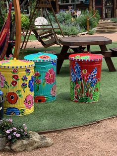 three brightly painted trash cans sitting on top of a grass covered field next to a picnic table