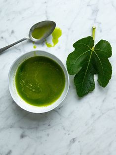 a white bowl filled with green liquid next to a leaf on a marble counter top