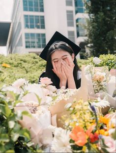 a graduate covers her face as she lays on the ground surrounded by flowers