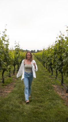 a woman walking down a path between rows of green grapes in a vineyard with trees behind her