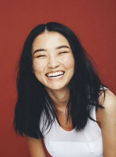 a woman smiling with her eyes closed in front of a red wall and wearing a white tank top