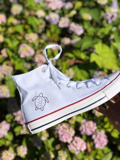 someone is holding up a white shoe with a turtle embroidered on the side, and pink flowers in the background