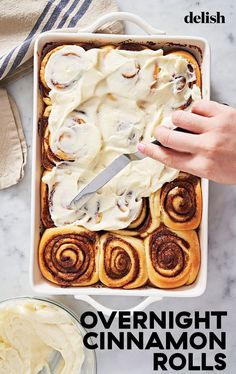 someone is spreading cream over cinnamon rolls in a baking dish with a knife on the side