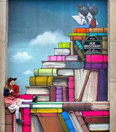 a woman sitting on top of a pile of books next to a mural painted on the side of a building