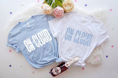 two t - shirts with the words on cloud in white and light blue, next to a bottle of booze