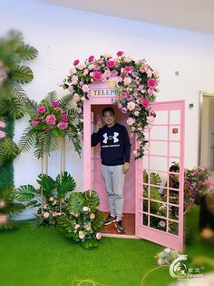 a man standing in a pink phone booth with flowers on the wall and greenery around it