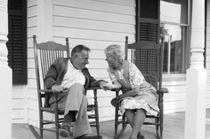 an old man and woman sitting on rocking chairs talking to each other in front of a house