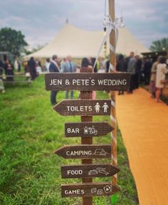 a wooden sign pointing in different directions at an outdoor wedding venue with people standing around