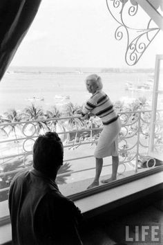 an older woman standing in front of a window looking out at the ocean and beach