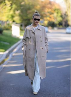 a woman walking down the street wearing a long coat and white pants with her hair in a bun