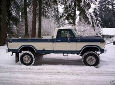 a blue and white truck parked in the snow next to some trees with lots of snow on it