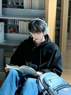 a young man sitting in front of a bookshelf reading a book with headphones on