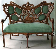 an ornate wooden bench with green velvet upholstered seat and back cushions, in front of a white wall