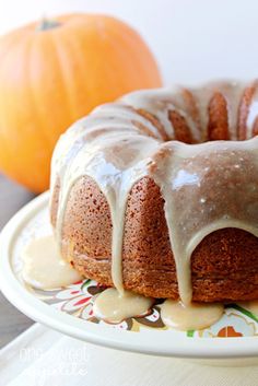 a bundt cake with icing sitting on a plate next to an orange pumpkin