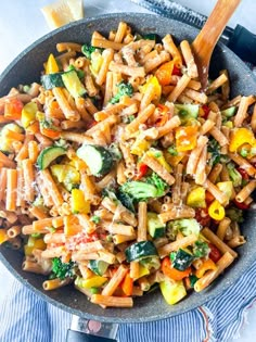 a pan filled with pasta and vegetables on top of a blue towel next to a wooden spoon