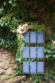 a blue sign with writing on it sitting next to some flowers and greenery in front of a stone wall