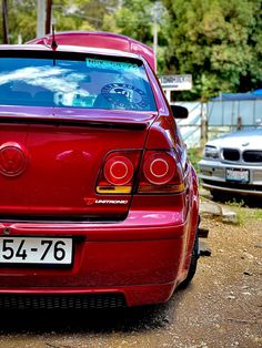 the back end of a red car parked in front of other cars on a dirt road